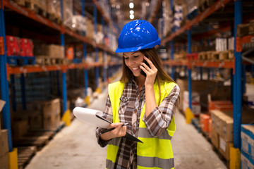 Warehouse worker with hardhat having conversation on the phone and holding checklist in distribution warehouse facility.