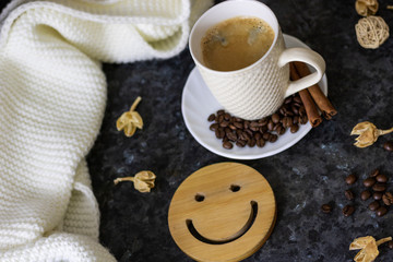 A cup of coffee is the key to a good mood. Wooden smiley on a dark, black, textural background. On the table there is a white, cream scarf and a white cup with espresso, coffee beans and cinnamon