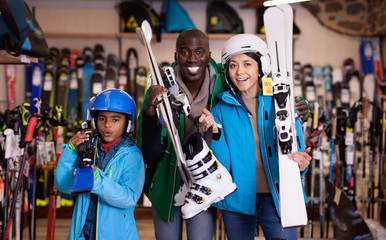 Fototapeta na wymiar Smiling young African man and European woman with preteen son standing with purchased ski equipment in shop