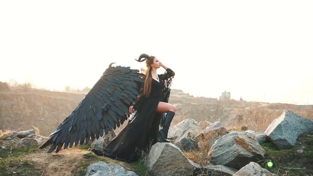 girl long vintage dress with flying train fashion model posing. Summer nature montains stone, sexu woman large feather wings horns on her head, image demon fallen black angel, creative colors sunlight