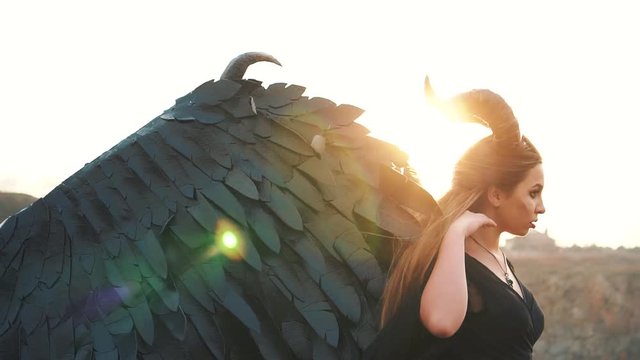 lady dark vintage dress with huge heavy strong wings behind her back removes her long hair from her face with gentle hand, demon with awesome black horns, portrait with creative colors, sun rays
