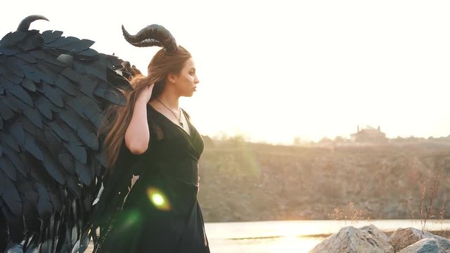 angel of death and war, girl in black long vintage dress with lace sleeves alone beside large lake, lady with strong feather wings down on ground and horns, portrait with creative colors, sun rays