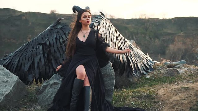mysterious lady in black light lace dress with long train, girl with open leg in leather boots, dark horned angel from heaven, messenger of death with large wings on edge sitting on stone rock