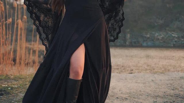 goddess of war and blood feud, fallen angel in long vintage black dress with loose lace sleeves with big horns on head raises her arms to sides and enjoys wind during sunset, portrait creative colors