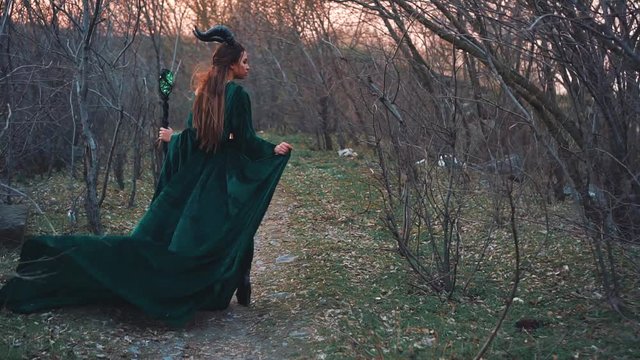 ancient spirit of forest, lady in a long emerald velor dress with long flying sleeves and hem, evil nymph goes to punish people for dirty nature, girl with magic stick in her hands and horns on head