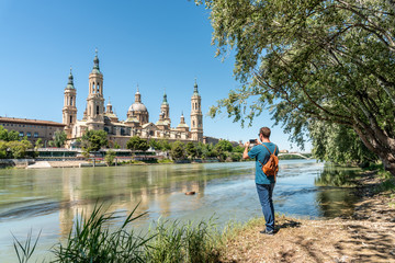Fototapeta premium Male tourist standing at the Ebro river bank and taking a photo of El Pilar in sunny day. 
