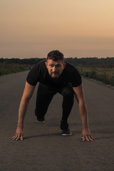 Mature fitness coach or runner in black sport joggers and tee stands in getting ready pose on a free countryside road in the evening time