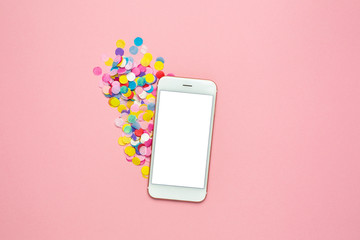 Mobile phone mock up and multicolored confetti on pink pastel table in flat lay style. Holiday mood.