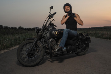 Obraz na płótnie Canvas Portrait of stylish manly biker on all black retro motorcycle with satisfied smile on face on free road with sunset and forest on background