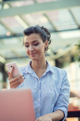 Portrait of a playful young girl taking selfie with mobile phone while sitting with laptop computer at a cafe outdoors