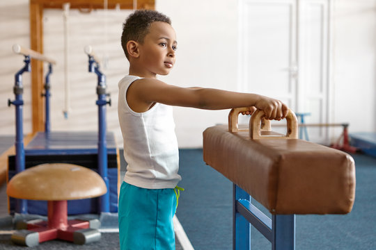 Picture of adorable happy dark skinned atheltic child in blue shorts and white t-shirt posing at gym with gymnastics equipment in background, holding hands on handles, going to perform routine