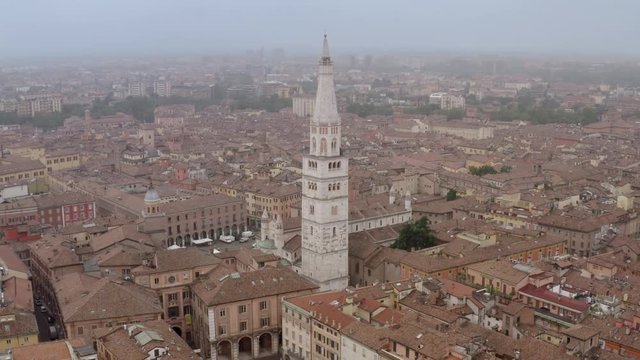Panoramic drone shot of the Ghirlandina tower, Modena, Italy, in the middle of the city, surrounded by buildings and streets, on a cloudy and foggy day