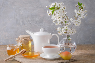 A cup of tea, a white kettle, pears, jars and a saucer with honey, a bouquet of cherry blossoms on the table. Morning breakfast.