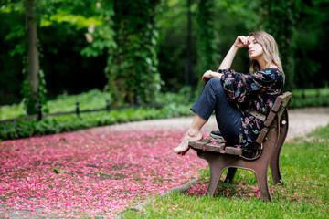 barefoot woman is sitting on a bench at alley with blossom trees in springtime.