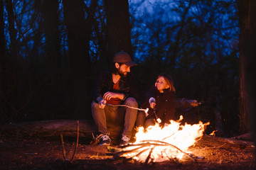 The father and his son sitting on the logs in the forest in front of a fire and roasting...