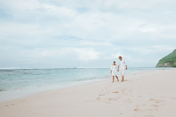 Girl and man in white clothes walking on white beach in Bali and holding hands