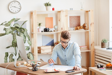 Fototapeta premium Serious skilled young male engineer in eyeglasses sitting at desk and using pair of compasses while measuring lines on blueprint