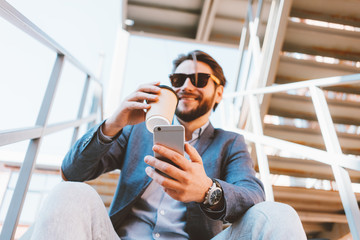 Young handsome businessman with stubble and sunglasses sitting on the stairs outside.  Drinking coffee to go and looking at mobile phone