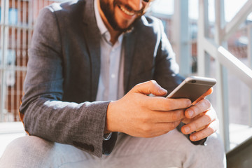 Young handsome businessman with stubble and sunglasses sitting on the stairs outside.  Smiling and using mobile phone, close-up