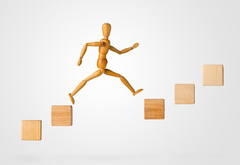 Wooden stick figure jumping from one wooden block on rising steps to the next - achievement, career...