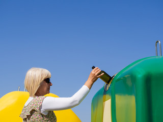A mature woman pulling a crystal bottle in a green bin for recycling glass with the spanish text "only glass containers"