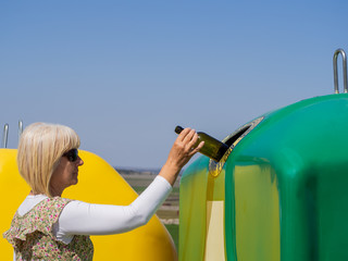 A mature woman pulling a crystal bottle in a green bin for recycling glass with the spanish text "only glass containers"