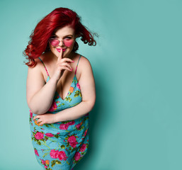 Overweight redhead lady plus-size woman in sunglasses shows Shhh gesture sign with her finger on lips and smiles enigmatically