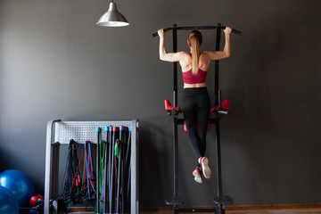 Young pretty muscular woman doing pull-up on horizontal bar. Stylish interior of modern fitness club with black walls. Concept of health and sport lifestyle. Athletic Body..