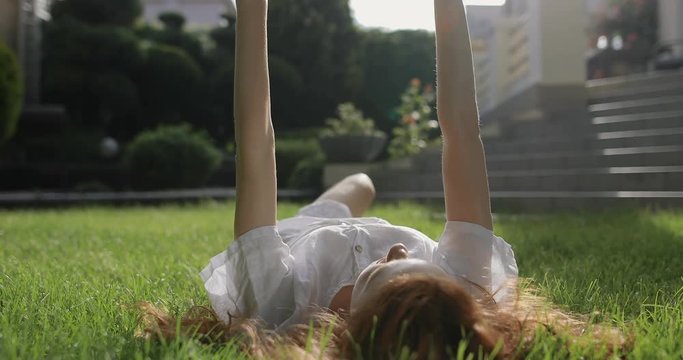 A young, attractive red-haired girl in a white dress lying on the grass near the house enjoying a wonderful warm summer day. A woman lies on her back and pulls her hands to the sun