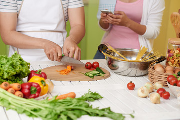 Young woman texting when her boyfriend cutting ingredients for sauce and boiling spaghetti
