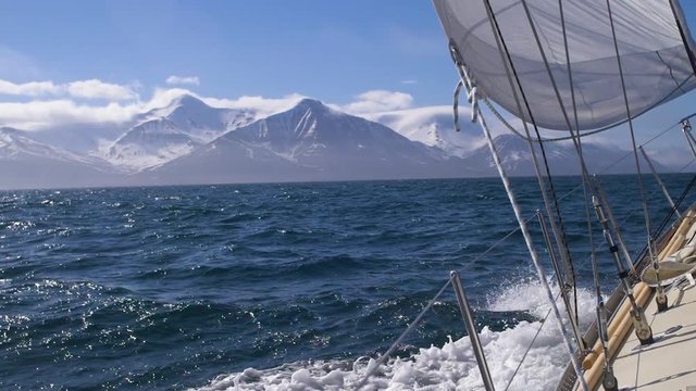 Sailing a yacht in Arctic with snow capped mountains - waves crash against hull in slow motion.