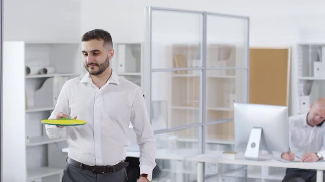 Thigh-up shot of male employee, wearing white shirt, enjoying active break in open space office, hitting ping pong ball with paddle repeatedly, while two colleagues are working on computers