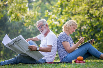 Happy senior couple relaxing sitting back to back in park reading newspaper and take write notes to Diary book  to together . old people in the summer park . Elderly resting .mature relationships . - Powered by Adobe