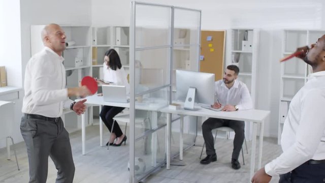 Medium shot of Afro-American and Caucasian male employees enjoying game of ping-pong during break, while female colleague stands up from desk and walks out with phone to shoulder and clipboard