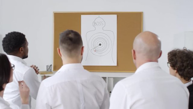 Chest-up rear shot of five excited office workers, wearing white shirts, playing funny teambuilding game and throwing darts at poster with target and word &ldquo;Boss&rdquo; on it, cheering, whooping and hugging