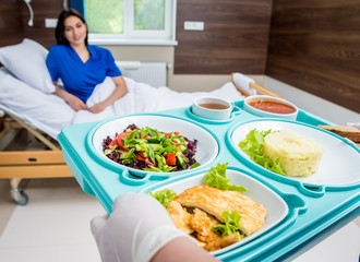 Nurse in medical coat is holding a tray with breakfast for the young female patient.