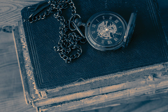 Antique pocket fob watch with a chain on a stack of old books. On a wooden background with colour toning. © Christian