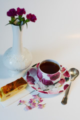 Still life of a morning cup of tea with sugar in a mug, a slice of appetizing cheese casserole with raisins and a miniature vase with three red small carnations on a white background