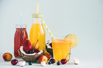 Yellow fruit juice in a bottle and in a glass with a straw, fresh fruit, coconut halves, apricots, fresh berries, cherry, lime, blueberries on a blue background. Copy space. Summer meal concept