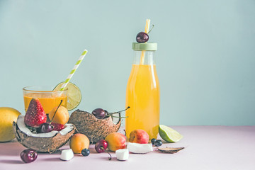Yellow fruit juice in a bottle and in a glass with a straw, fresh fruit, coconut halves, apricots, fresh berries, cherry, lime, blueberries on a blue background. Copy space. Summer meal concept