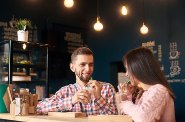 Young stylish happy couple sitting at cafe table, drinking coffee and looking at each other. Young man and woman at coffee shop. Romantic acquaintance concept. Creative design blue wall on background.