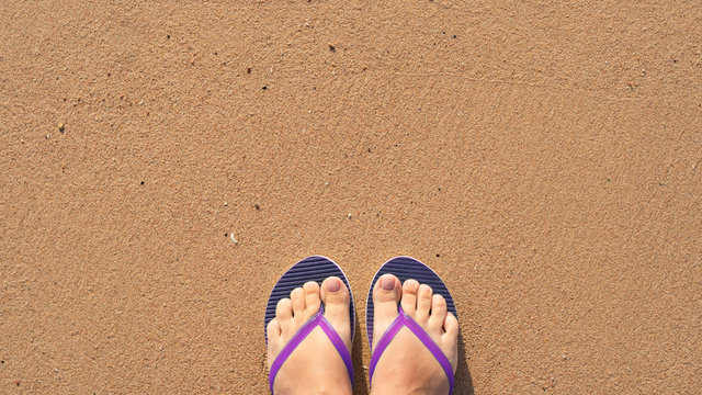 Point of view photography of beautiful female legs with natural pink nude look pedicure wearing rubber colorful flip flops standing alone at sandy summer sea beach. Happy holidays and travel concept.