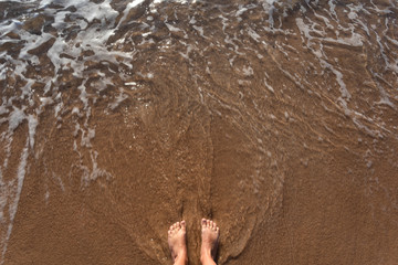 Top view of two female beautiful legs isolated at sandy seashore with splashing foamy warm sea water. Happy summer holidays and travelling concept. GHorizontal color image with copyspace.