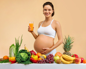 Pregnant woman with vegetables and fruits on the table, holds orange juice on beige background....