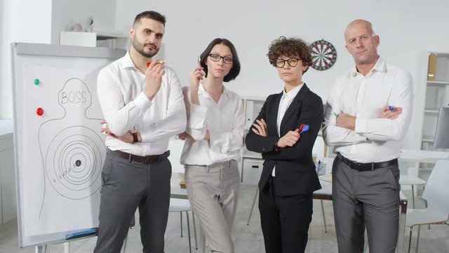 Dollying high-angle shot of four company employees, dressed in smart business outfits, posing together next to flipchart with target poster named &ldquo;boss&rdquo;, with arms folded on chest and holding darts