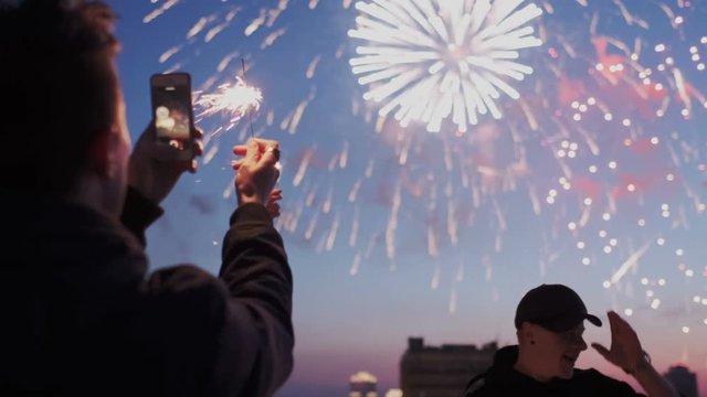 Young guy is holding a smartphone with sparklIng light and recording a video of fireworks on the rooftop. Friends smiling and having fun on the background. Holidays. Slow motion.