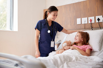 Female Nurse Visiting Girl Lying In Hospital Bed Hugging Teddy Bear