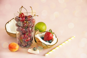Fruitorianism.Cherry berries in a transparent glass cup on a pink background next to coconut halves, strawberries, blueberries, lime, apricots. The concept of healthy eating. Fruit dessert. Copy space