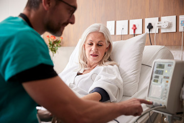 Male Nurse Taking Mature Female Patients Blood Pressure In Hospital Bed With Automated Machine