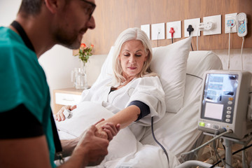Male Nurse Taking Mature Female Patients Blood Pressure In Hospital Bed With Automated Machine - obrazy, fototapety, plakaty